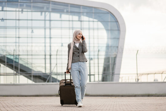 Portrait of happy muslim arab woman wearing a hijab and casual clothes, walking outdoors carrying a suitcase and talking phone, going to travel by airplane at modern airport.
