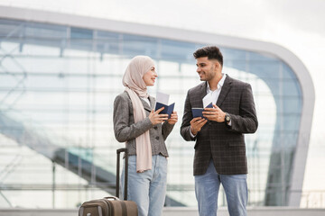 Fototapeta na wymiar Excited Muslim couple with suitcase, standing outdoors at airport and checking their flight schedule, looking at the ticket and smartphone. Business trip together. People, travel, technology concept