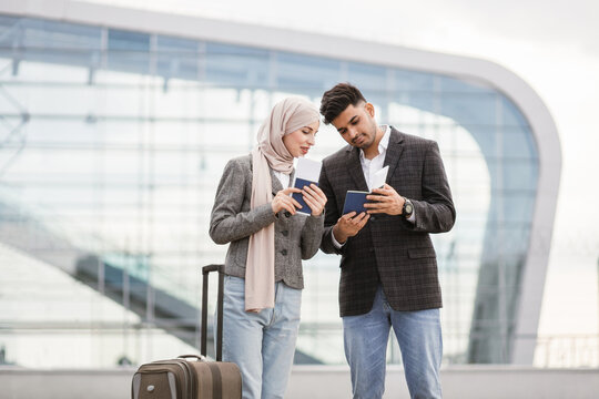 Excited Muslim couple with suitcase, standing outdoors at airport and checking their flight schedule, looking at the ticket and smartphone. Business trip together. People, travel, technology concept