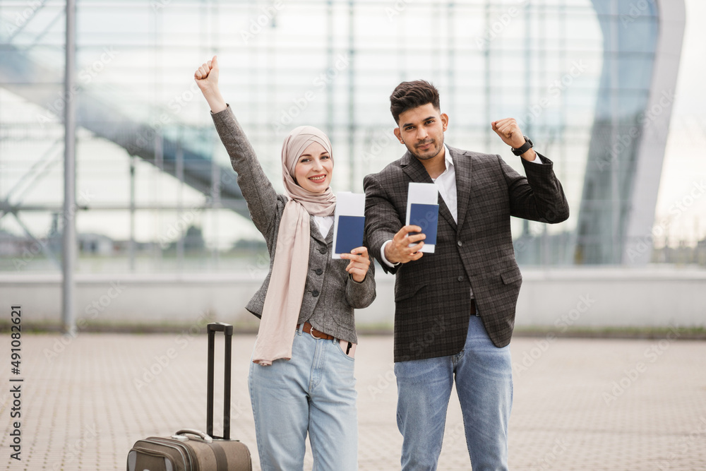 Wall mural smiling muslim woman in hijab and arab man, posing on camera outside modern airport terminal, demons