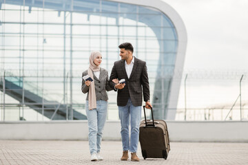 Portrait of happy muslim couple, woman in hijab and manin jacket, walking with passports and tickets outdoors carrying a suitcase and talking each other, going to travel by airplane at modern airport.
