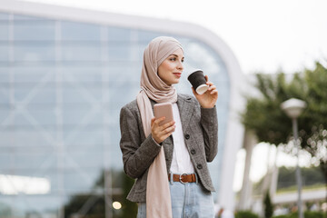 Pretty happy Muslim lady in hijab smiling at camera, standing outdoors with coffee to go and phone in hands. Likable Arab woman in headscarf walking on the street with take away coffee and phone