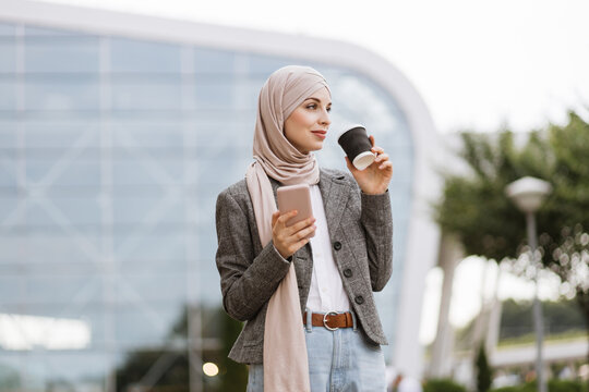 Pretty happy Muslim lady in hijab smiling at camera, standing outdoors with coffee to go and phone in hands. Likable Arab woman in headscarf walking on the street with take away coffee and phone
