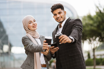 Smiling business couple, handsome Arab man and Muslim woman in hijab, standing outdoors in the street on coffee break, using phones and drinking take away coffee