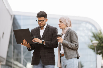 Fototapeta premium People and technologies concept. Attractive business couple of arab man and Muslim woman in smart casual style, standing outside office, working together on laptop, smiling and discussing