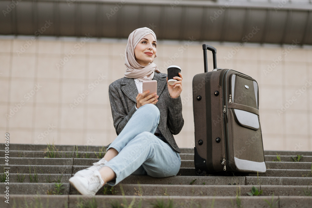 Wall mural young muslim woman in hijab dressed in jeans and jacket using phone and drinking take away coffee, w