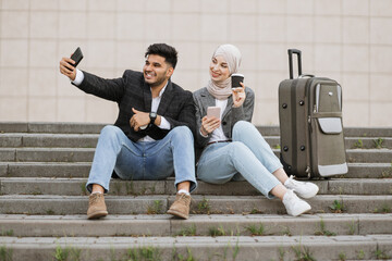 Pleasant smiling Indian man and pretty Muslim lady in hijab, sitting on the stairs with travel suitcase and and doing selfie. Business couple sitting on the stairs outside airport.