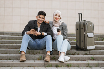 Two multiracial business people sitting at stairs with travel bag and looking at camera. Diverse colleagues ready for the trip, sitting with phones and coffee on strairs outside