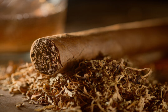 The cigar lies on a pile of tobacco and a glass of whiskey stands nearby.