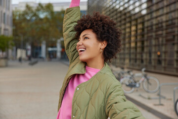 Outdoor shot of positive curly haired young woman dressed in jacket laughs positively enjoys...