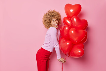 Sideways shot of overjoyed curly haired woman wears casual jumper and red trousers holds bunch of heart inflated balloons has fun during celebration isolated over pink background. Holidays concept