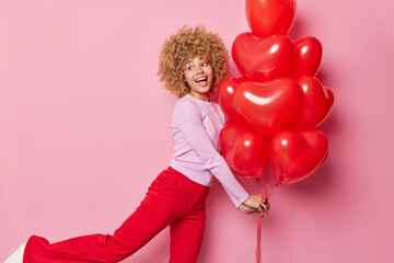 Cheerful woman with curly hair dances carefree holds bunch of heart balloons keeps mouth opened feels very glad wears casual jumper and trousers isolated over pink background. Happy Valentines Day