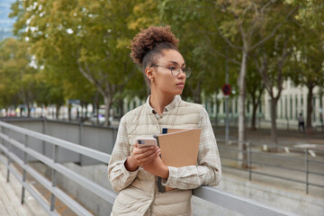 Outdoor shot of thoughtful curly haired woman uses mobile phone for chatting online holds notebooks poses in city against park wears round spectacles and vest has pensive expression. Lifestyle
