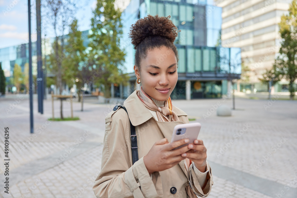 Wall mural Outdoor shot of curly haired female dressed in coat messages on smartphone while walks in city downloads music to playlist uses free internet connection sends sms in chat writes new travel story