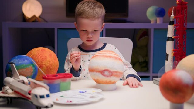 Blond boy paint planet solar system Jupiter with colorful paint sitting home table in evening, planet solar system, spaceships and space shuttle from constructor around. Cosmonautics Day on April 12.