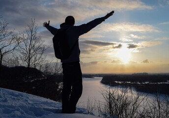 Silhouette of a man with outstretched arms against the background of a winter sunset and a frozen river