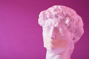 copy of the head of an antique statue of David with a taped mouth in pink neon light on a purple background