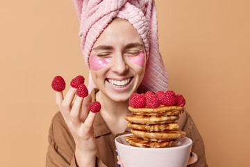 Sincere cheerful young woman smiles gladfully keeps eyes closed eats delicious pancakes with syrup has raspberries on fingers wears wrapped towel and pajama applies hydrogel patches poses indoor