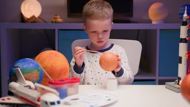 Blond boy paint planet solar system Venus with colorful paint sitting home table in evening, planet solar system, spaceships and space shuttle from constructor around. Cosmonautics Day on April 12.