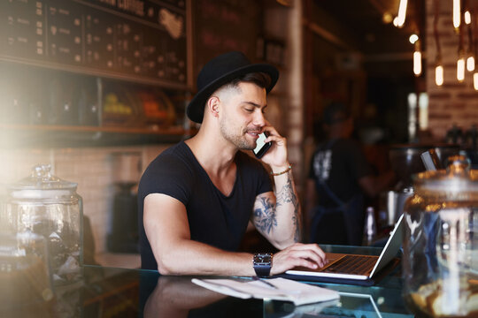 Busy behind the bar. Shot of a young man using a phone and laptop while working in a coffee shop.