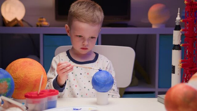 Blond boy paint planet solar system Pluto with colorful paint sitting home table in evening, planet solar system, spaceships and space shuttle from constructor around. Cosmonautics Day on April 12.