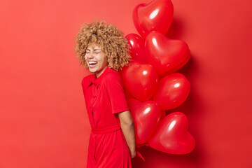 Overjoyed curly haired young woman laughs out happily holds big bunch of heart balloons behind back prepares surprise for lover on Valentines Day wears dress isolated over vivid red background.