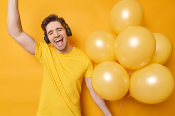 Horizontal shot of happy handsome man holds bunch of inflated balloons has fun dances carefree...