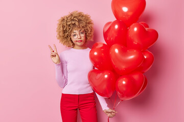 Sad serious curly woman tries to entertain someone makes peace gesture holds bunch of heart inflated balloons wears spectacles jumper and trousers isolated over pink background. Special occasion