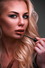 Obraz premium Portrait of young blonde woman painting her lips with lipstick on gray background