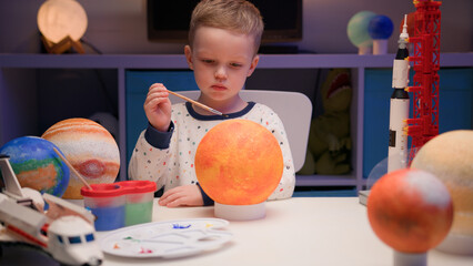 Blond boy draws sun star with multicolored paint, sitting at home table in evening, planet of solar system, spaceships and space shuttle from constructor around. Cosmonautics Day is April 12.