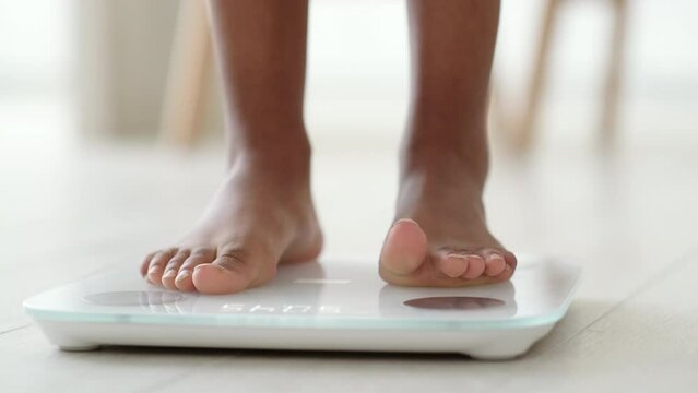 Legs of black girl standing on scale to measure weight. African American Female bare feet with weight scale at home. dieting, control and measuring 