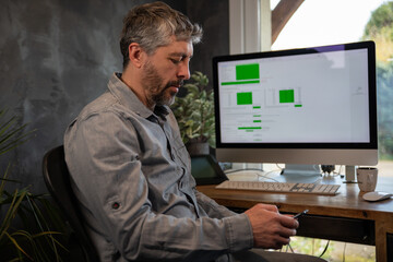 homme de 50 ans en télétravail dans son bureau devant son ordinateur. il consulte son téléphone...
