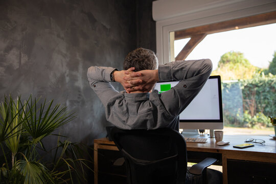 vue de dos d'un homme qui travaille chez lui en t&eacute;l&eacute;travail et qui se relaxe sur son fauteuil de bureau en mettant ses mains derri&egrave;re sa t&ecirc;te