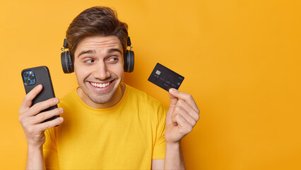 Optimistic carefree handsome man holds modern smartphone and banking card uses application on cellphone for online payment isolated over yellow background blank space for your promotional content.