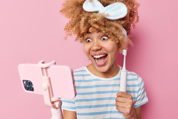 Pretty young woman with curly combed hair holds electric toothbrush shoots video holds mobile phone on stick wears headband and striped t shirt isolated over pink background. Teeth care concept