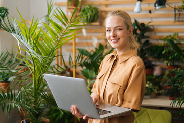 Portrait of cheerful businesswoman floral store owner standing with laptop holding in hand among...