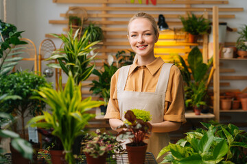 Medium shot portrait of smiling female florist in apron holding in hands pot with Soleirolia plant standing in floral shop, looking at camera. Woman gardener posing with houseplants at home.