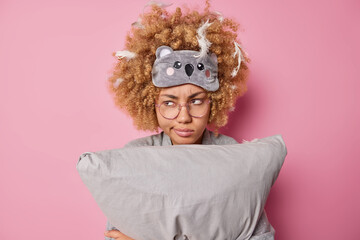 Serious displeased curly haired woman holds pillow looks discontent away wears sleepmask on forehead has feathers stuck in curly hair poses against pink background expresses negative emotions