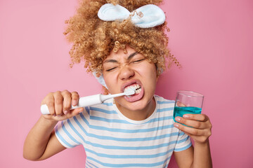Indoor shot of curly haired young woman brushes teeth with whitening toothpaste uses electric...