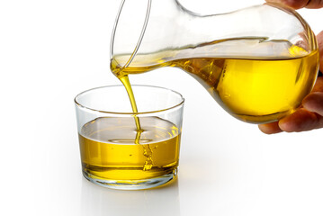 Extra virgin olive oil being poured from decanter into glass cup on white background