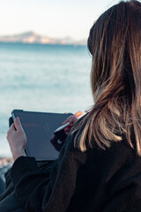 Back view of young girl drawing on tablet with pen by sea. Her face is not see. Unrecognizable person. Creativity and design concept. Vertical photo.	