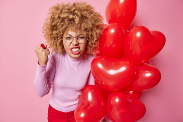 Irritated woman clenches teeth and fist feels annoyed holds bunch of heart balloons comes on party wears spectacles and casual clothes isolated over pink background. Holidays and people concept