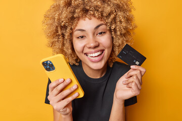 Happy curly female shopper enjoys easy paying for goods online smiles broadly shows white teeth...