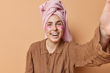 Positive young woman dressed in pajama towel wrapped on head giggles positively takes selfie with unknown device applies hydrogel patches enjoys starting of new day isolated over beige background.