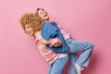 Two funny beautiful women give piggy back to each other have fun and laugh happily dressed in...