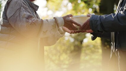 Poor homeless elderly person is being given a bowl of warm meal by a caucasian volunteer. Outdoor shot. High quality 4k footage