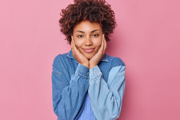 Portrait of gentle cute woman with curly hair keeps hands on cheeks smiles pleasantly looks directly at camera wears casual shirt poses against pink background. Human face expressions concept - obrazy, fototapety, plakaty