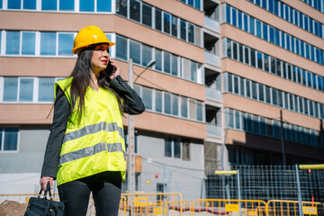 Adult ethnic female engineer with briefcase talking on smartphone near building