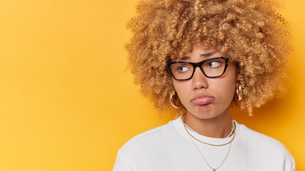 Horizontal shot of discontent woman with curly hair purses lips looks sadly away expresses negative emotions has troubles isolated over yellow background blank space for your promotional content