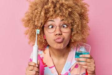 Photo of curly haired young woman makes funny grimace pouts lips holds electric brush and glass of mouthwash takes care of dental hygiene isolated over pink background. Morning routine concept